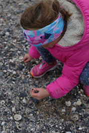 Young girl in a pink coat wearing the Beach Bounty Infinity Band in pinks, blues and navy as she kneels down to collect shells at the beach.