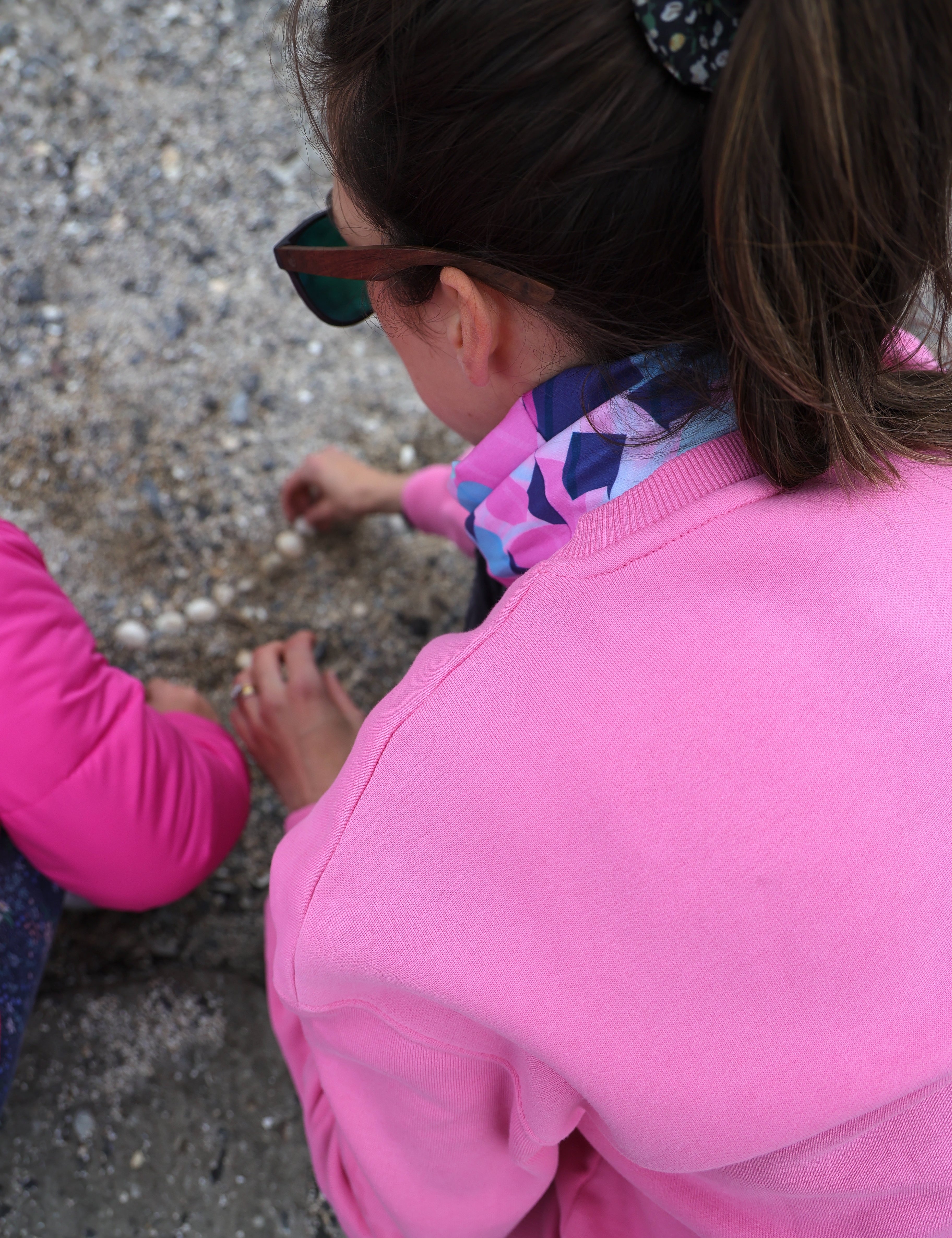 Lady wearing pink sweatshirt and the pink, navy and turquoise blue Infinity Band or snood around her neck while collecting shells at the beach with a young girl in a pink coat.